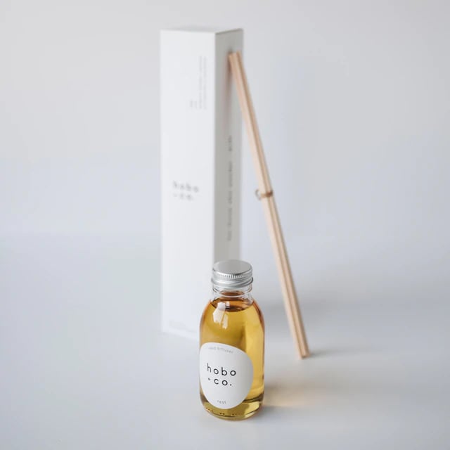 Kleine Flasche mit Duftöl von hobo + co. und Holzstäbchen vor einer weißen Verpackung auf einem weißen Hintergrund.