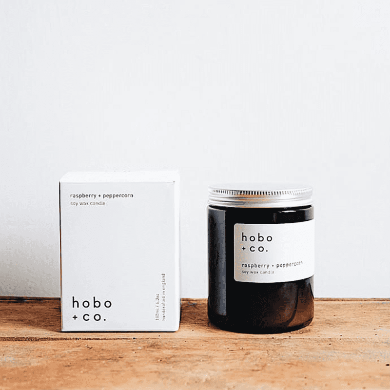 Hobo + Co. Sojawachskerze mit Himbeer-Pfefferkorn-Duft, 180ml, neben der Verpackung auf einem Holztisch.