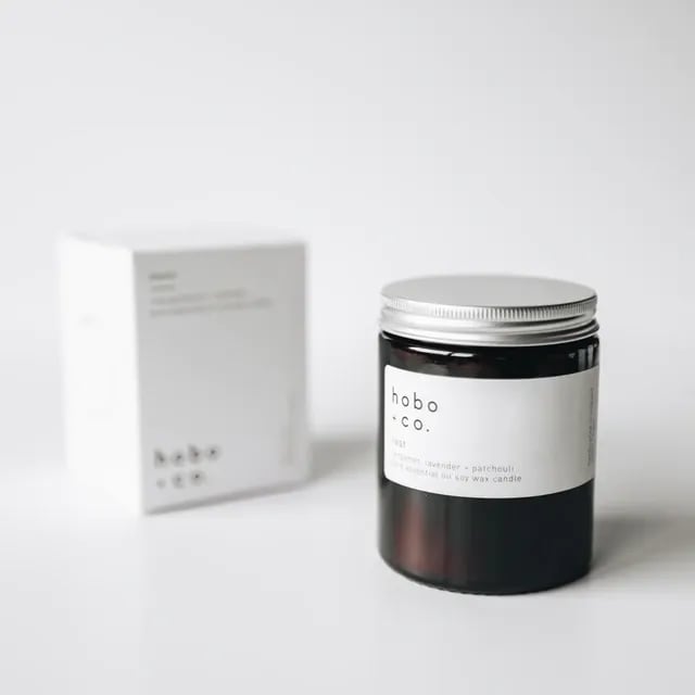 Eine schwarze Duftkerze von hobo + co. mit weißem Etikett und silbernem Deckel, daneben die passende Verpackung.