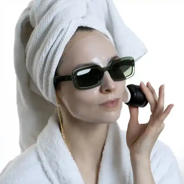 Frau mit Handtuch auf dem Kopf und Sonnenbrille trägt Gesichtsmaske auf.
